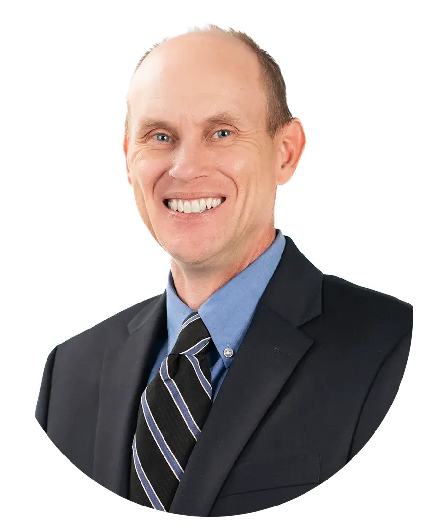 A middle-aged man with short light hair, wearing a dark suit, blue shirt, and striped tie, smiles at the camera against a white background.