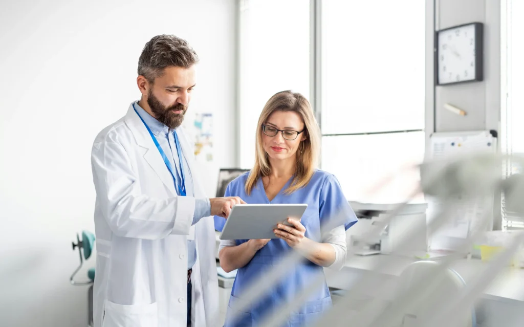 A male doctor and a female nurse stand together in a medical office, reviewing information on a digital tablet.