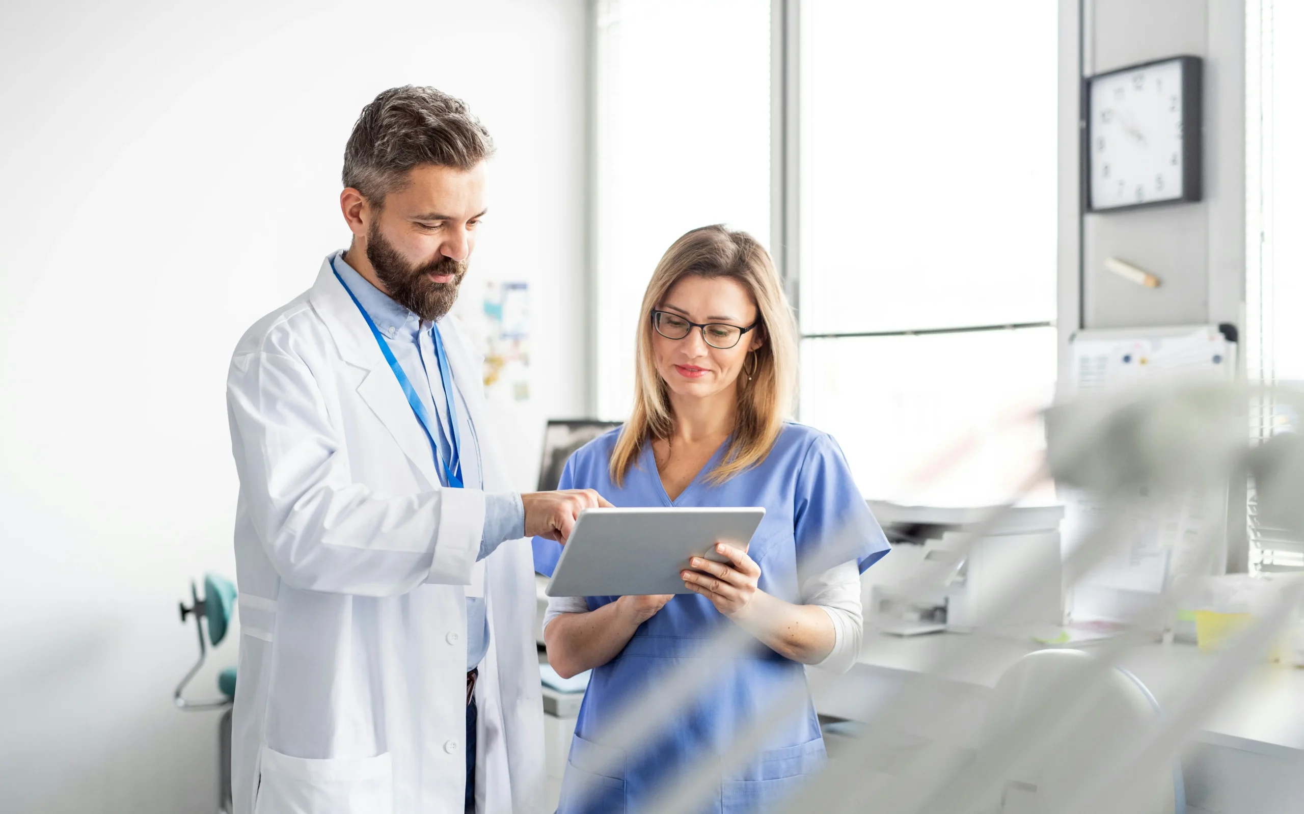 A male doctor and a female nurse stand together in a medical office, reviewing information on a digital tablet.