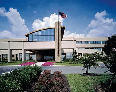 A modern, multi-story building with large windows and an American flag on a flagpole, surrounded by landscaped greenery and flowerbeds under a partly cloudy sky.