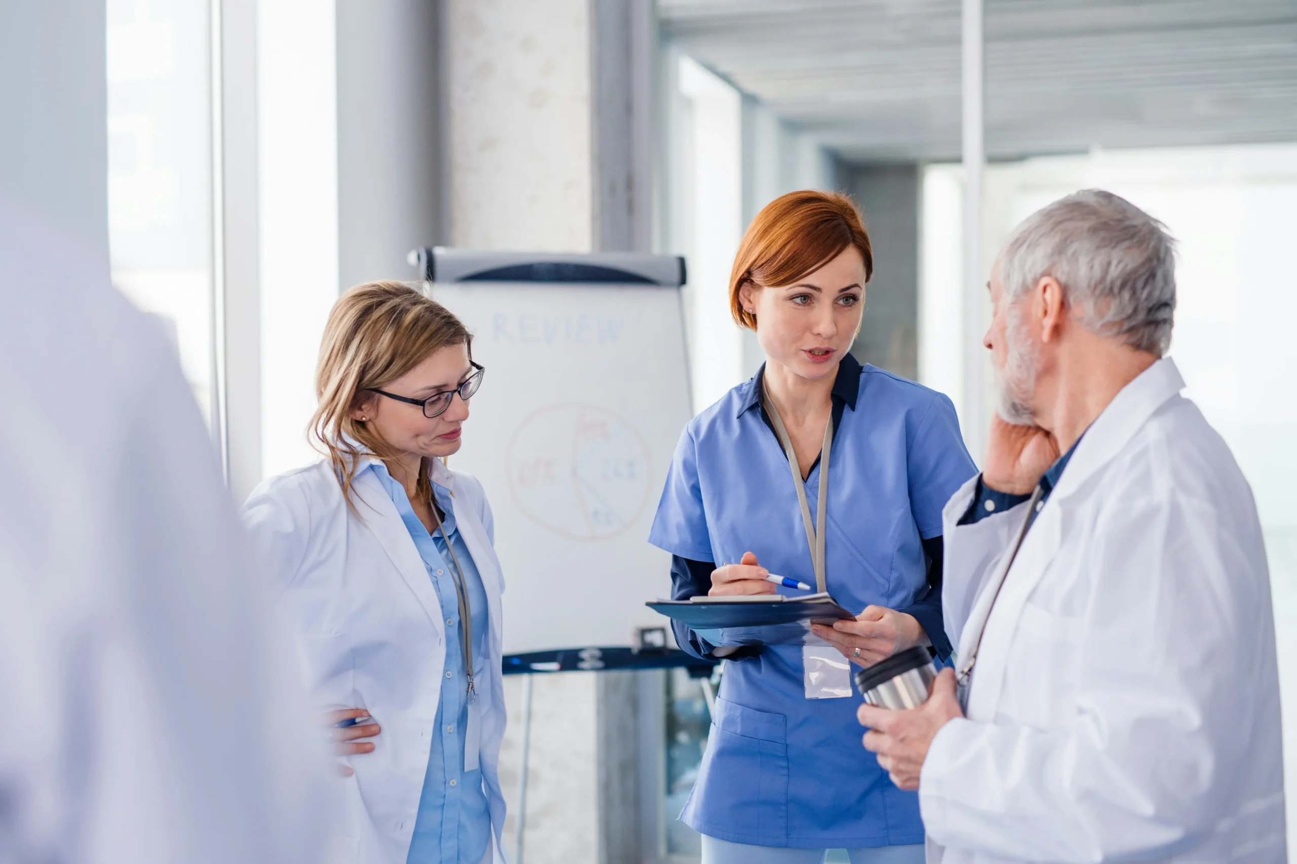 Three medical professionals in discussion near a flip chart; one holds a tablet, another a mug, all wearing scrubs or lab coats.