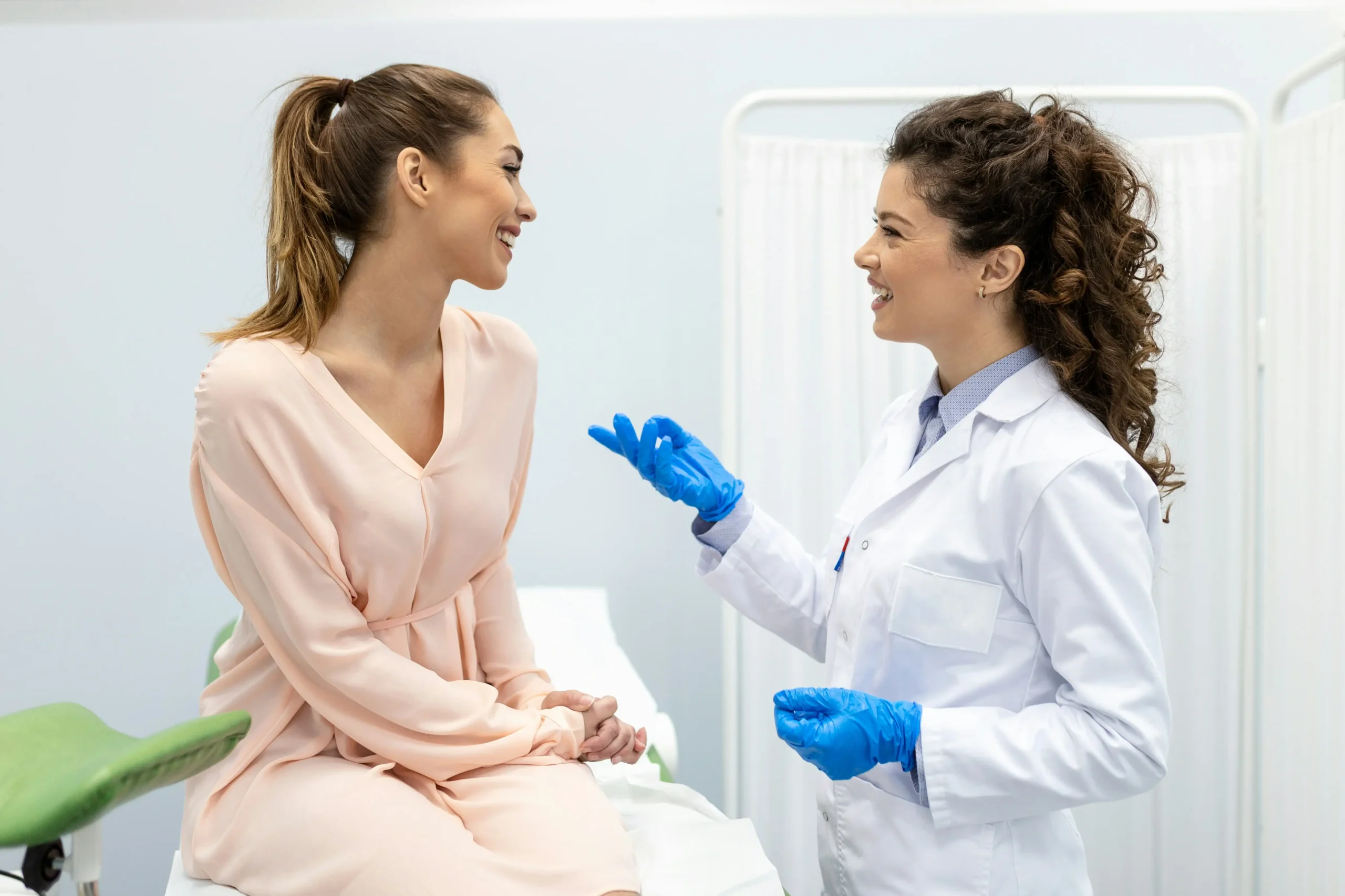 A doctor wearing blue gloves talks to a seated female patient in a medical exam room.