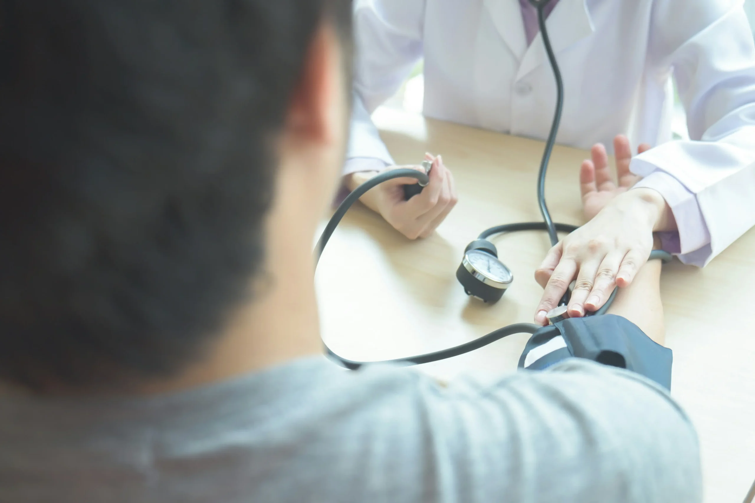 A healthcare professional measures a person's blood pressure using a sphygmomanometer and stethoscope at a desk.