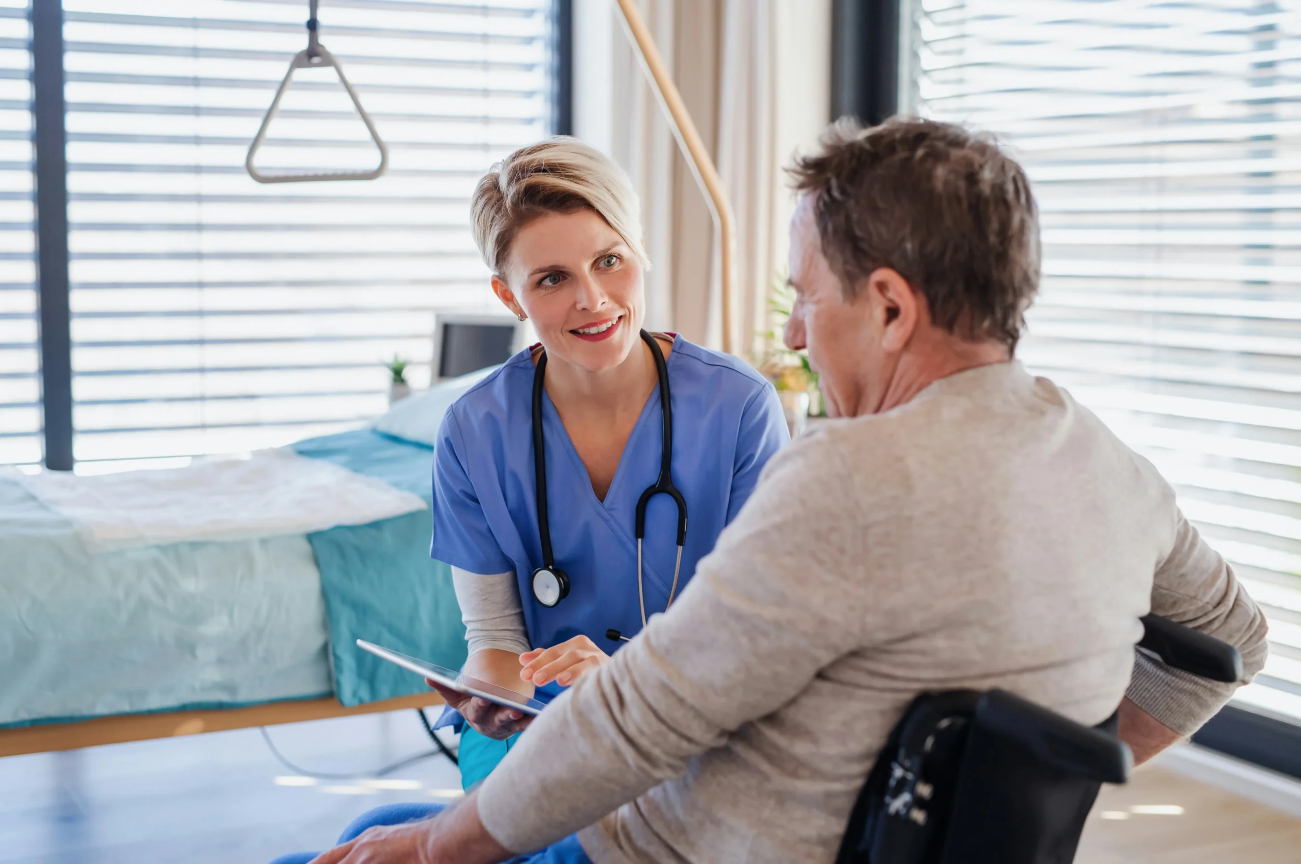 A nurse in blue scrubs holds a tablet and talks to a man in a wheelchair in a medical room with a hospital bed in the background.