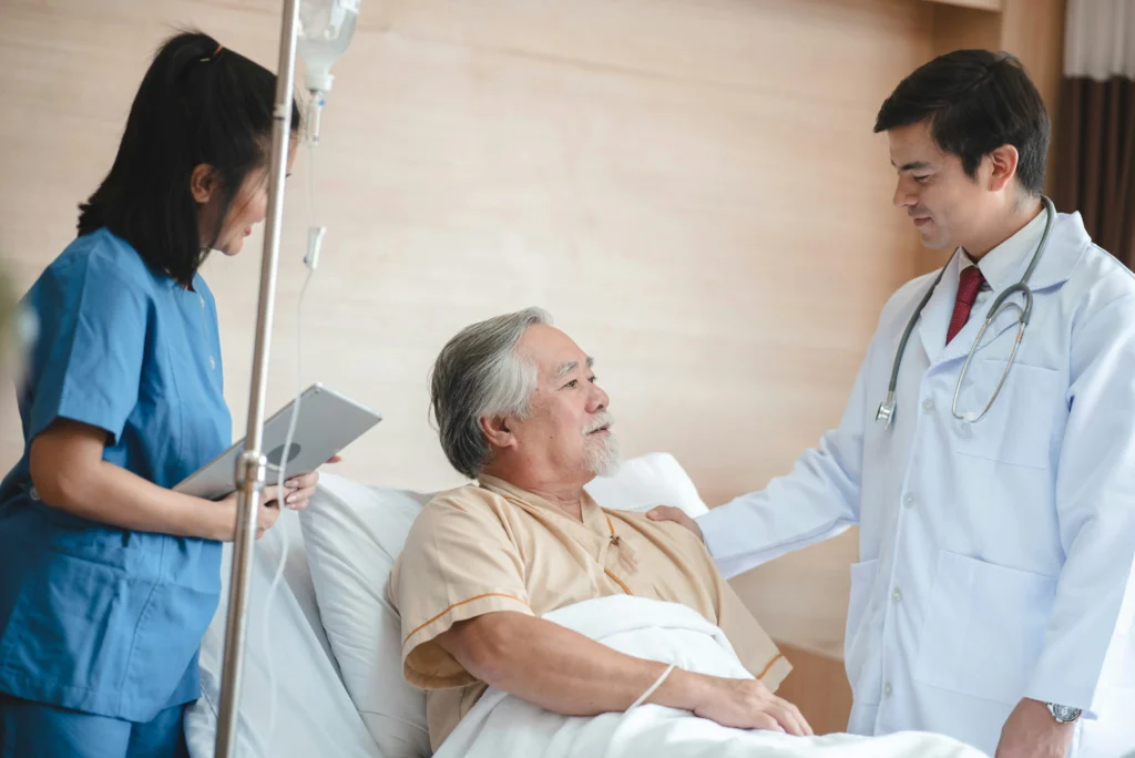 A doctor and nurse speak with an elderly male patient who is lying in a hospital bed, with an IV stand nearby.