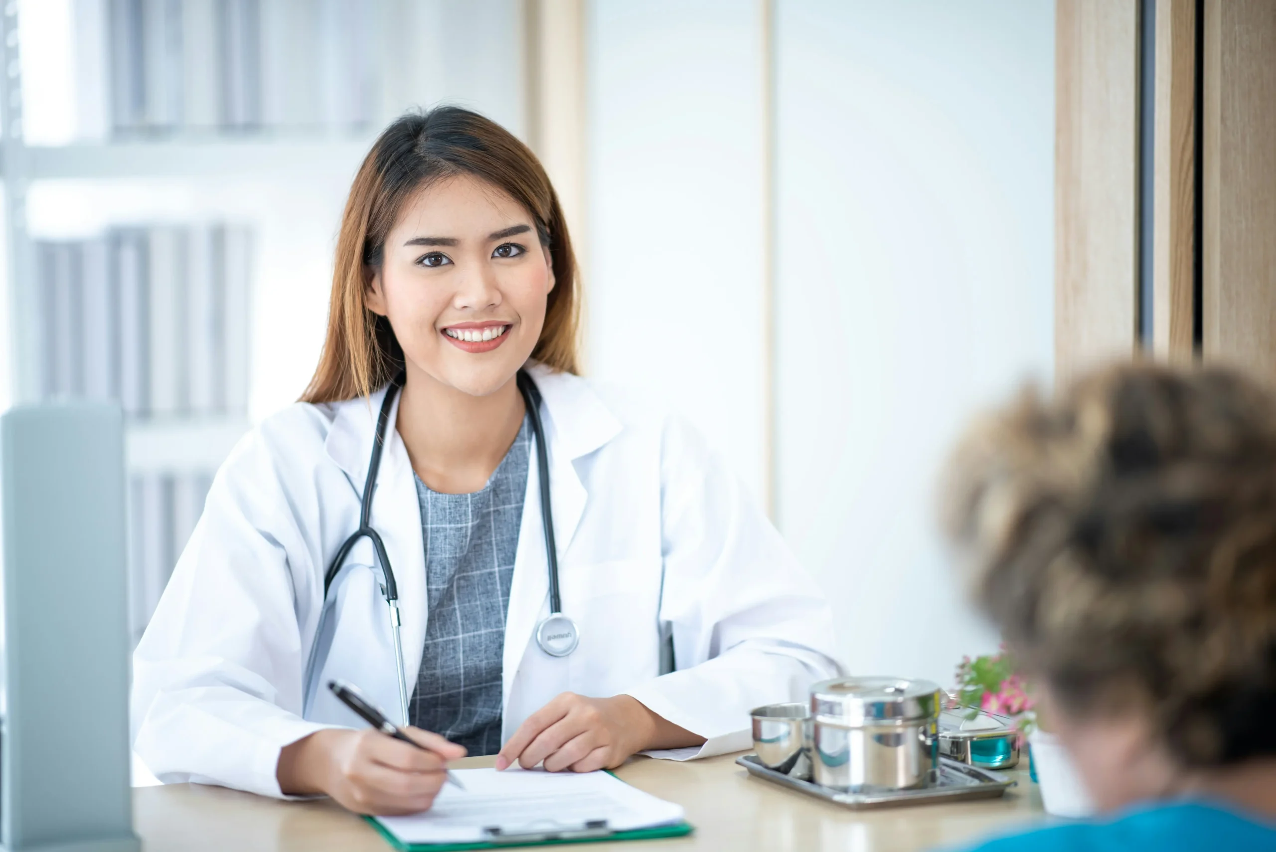 A doctor wearing a white coat and stethoscope sits at a desk, smiling, and writing on a clipboard while talking to a patient.