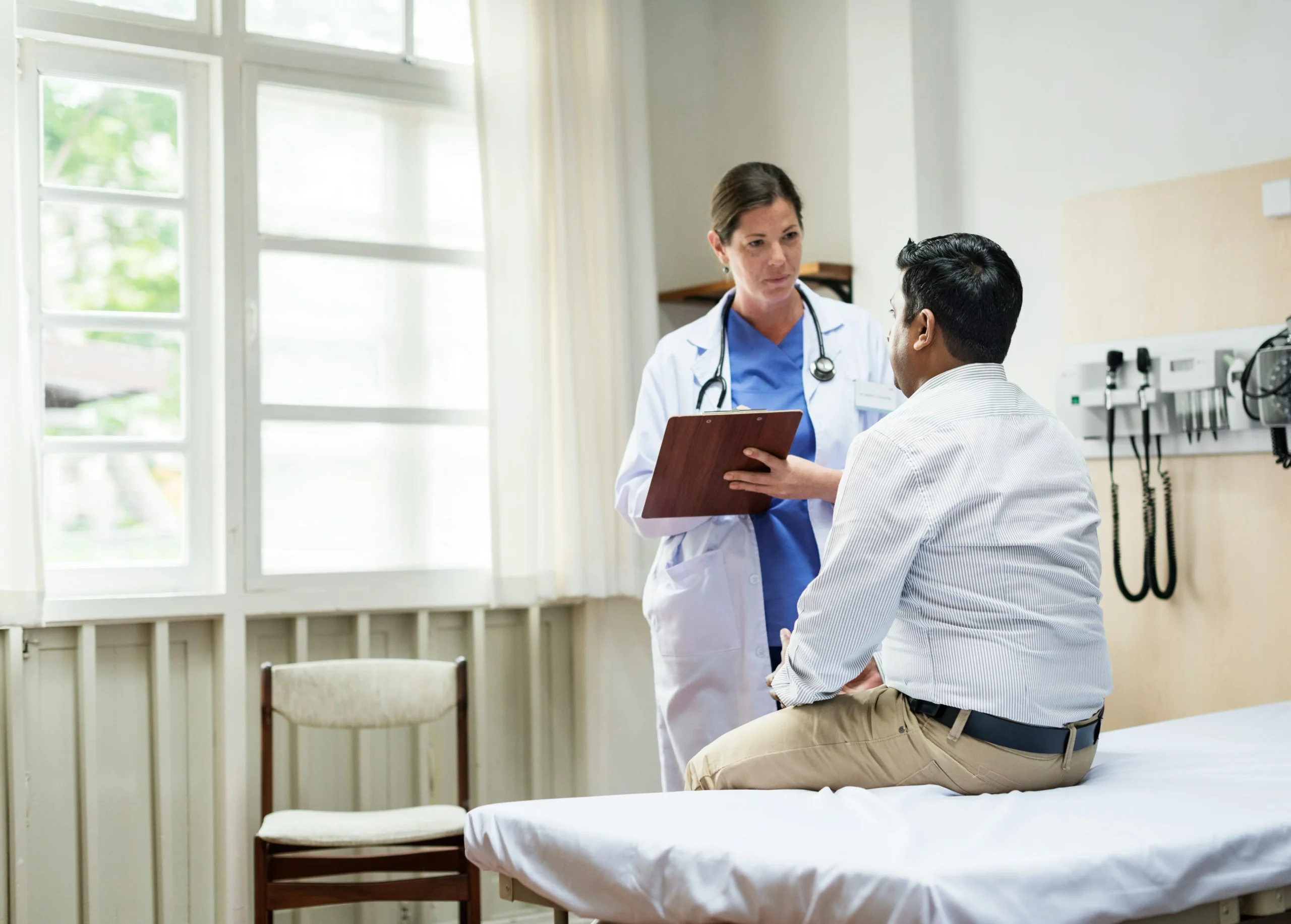 A doctor holding a clipboard talks to a patient sitting on an exam table in a medical office with natural light from a window.