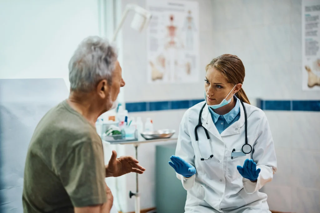 A female doctor wearing a white coat and gloves speaks with an older male patient in a medical office.