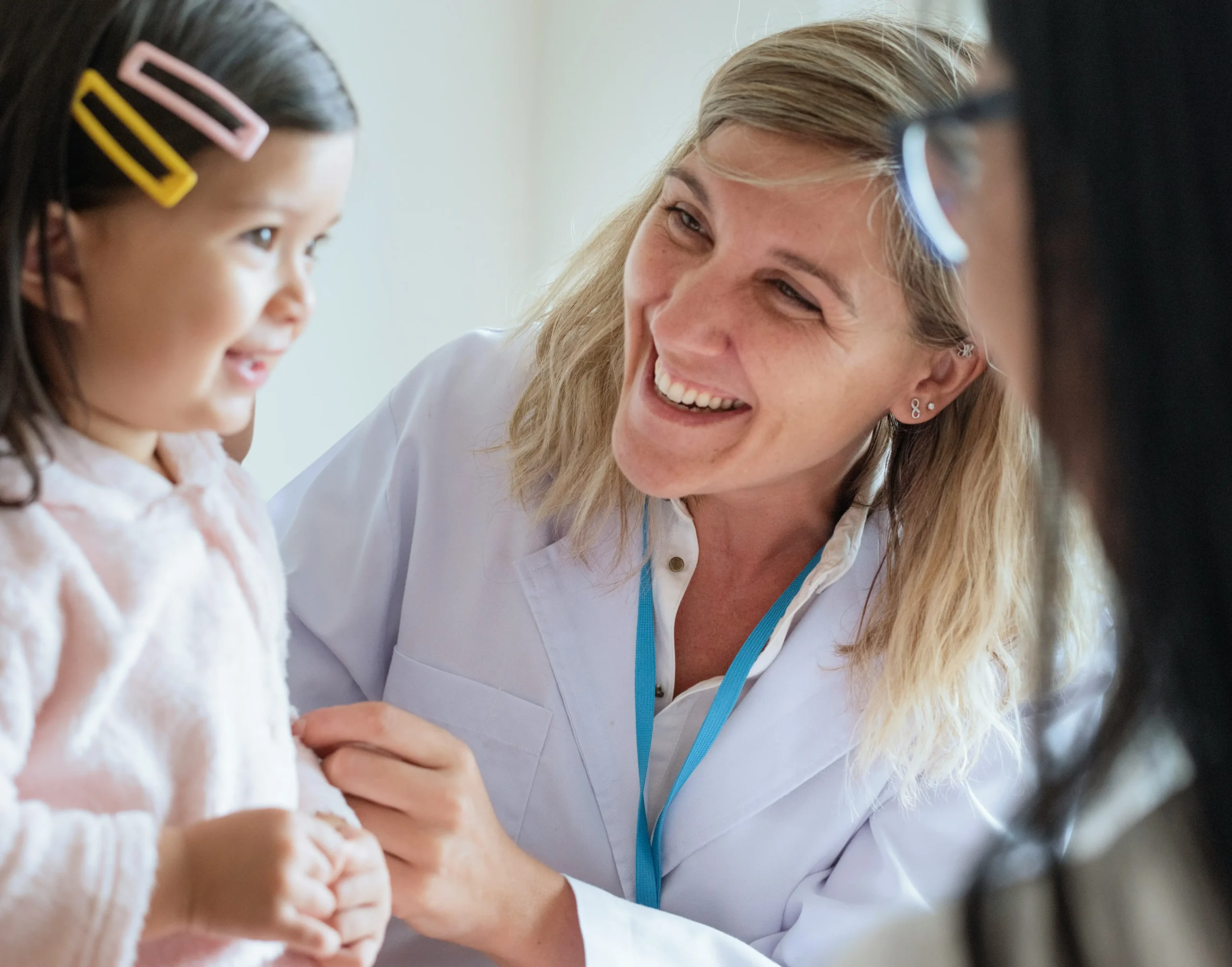 A female healthcare professional in a white coat smiles at a young girl with hair clips, while another adult is partially visible in the foreground.